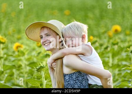 Glücklich lachende Mutter geben Kleinkind Sohn Huckepack Fahrt auf dem Hintergrund der grünen blühenden Sonnenblumen Feld. Stockfoto