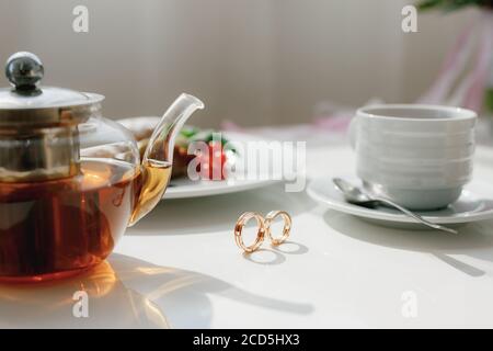 Hochzeit Goldringe auf einem Tisch mit einer vollen Teekanne und eine Tasse mit einem Löffel auf einer Untertasse. Stockfoto