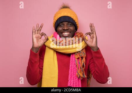 Erfolgreiche aufgeregt jungen afrikanischen Mann in mehreren Hut und Schal zeigt ok Geste. Stockfoto