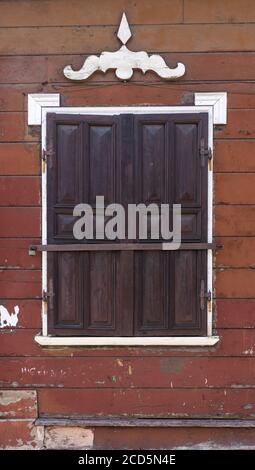 Details der traditionellen Architektur. Braune Holzwand mit Fenster geschlossen mit hölzernen Fensterläden mit Metallbeschlägen und weißen dekorativen Details. Stockfoto