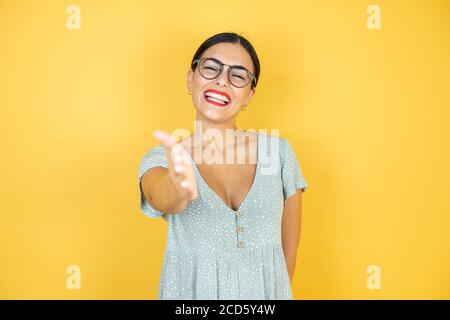 Junge schöne Frau trägt eine Brille, die über einem isolierten gelben Hintergrund steht Lächelnd und freundlich, mit Handschlag als Begrüßung und Begrüßung Stockfoto