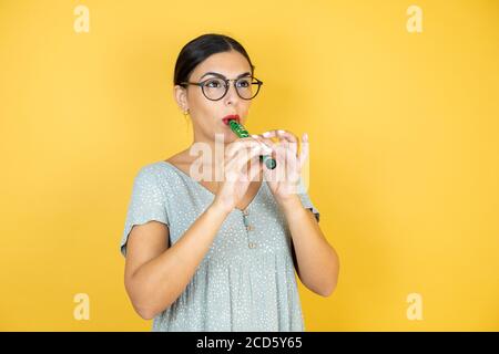Junge schöne Frau trägt eine Brille, die über einem isolierten gelben Hintergrund steht Flöte spielen Stockfoto