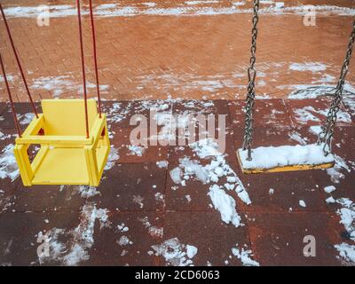 Zwei gelbe Holzschaukeln über nassen roten Pflasterplatten mit Die Reste von aufgetautem Schnee Stockfoto