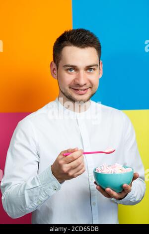 Ein Mann auf gelbem, blauem, orangefarbenem, rosa Hintergrund in einem weißen Hemd mit einem türkisfarbenen Teller Marshmallows und einem rosa Löffel. Der Typ mit einem Lächeln. Ein im Stockfoto