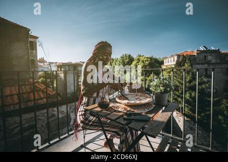 Eine entzückende junge schwarze Frau ist mit Mittagessen und Schneiden In Stücke leckere Pizza mit Käse und Speck im Sitzen Auf einem Balkon Stockfoto