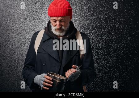 Älterer graubärtiger Mann mit eiserner Pfanne und Suppenkelle. Versuchen Sie, etwas Essen auf dem Boden der Pfanne zu finden. Obdachloser, der unter Regen steht Stockfoto
