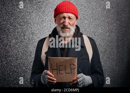 Älterer Bettler mit grauem Bartstand, der jedem, der um Hilfe bittet, ein Schild zeigt. Mann in Straßenkleidung Stockfoto