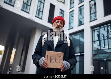Armer Mann hält Hilfe Schild aus Pappe gemacht, stehen im Zentrum der Stadt, große schöne Geschäftsgebäude hinter ihm. Stockfoto