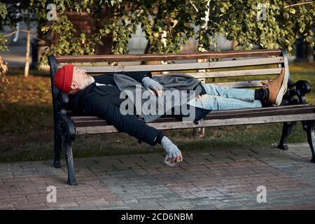 Obdachloser, der tagsüber auf der Bank im Park schläft. Reifer graubärtiger Mann in Straßenkleidung. Seitenansicht Stockfoto