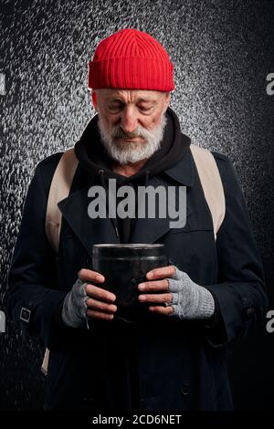 Bärtiger alter Mann, der schwarze Straßenkleidung trägt und einen Eisenbecher für Geld hält, um Hilfe zu erhalten. Trauriger Obdachloser unter starkem Regen. Stockfoto