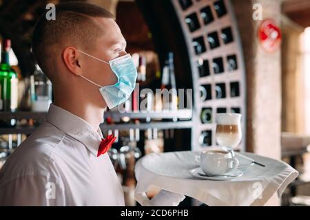 Ein europäisch aussehender Kellner in einer medizinischen Maske serviert Latte-Kaffee. Stockfoto