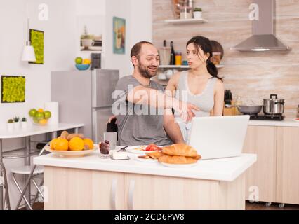 Ehemann lachend über die Frau, die ihr ein Bild im Laptop zeigt. Argument, Konflikt, verzweifelte Probleme streiten und Konflikt in der Ehe, traurige Emotionen und verzweifelte, unglückliche Paar Stockfoto