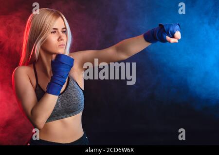 Junge attraktive Kickboxen weibliche Kämpfer mit blonden Haaren üben Schläge. Kickboxer in blauen Bandagen werfen Schläge für den Kampf. Dunkel verqualmt Stockfoto