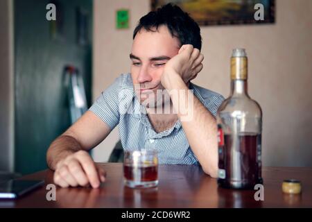 Trauriger, trauriger junger Alkoholiker sitzt mit einer Flasche Rum am Tisch. Das Gesicht eines betrunkenen Mannes. Dumme Emotionen. Depression und Alkoholismus. Stockfoto