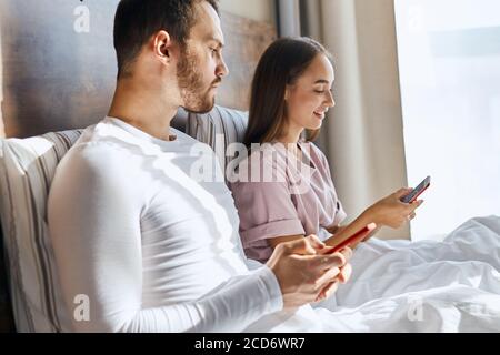 Fröhliche hübsche Frau sitzt auf dem Bett mit Handy in der Hand, gerne SMS mit Freunden im Chat, seriöse unrasiert Mann sitzt in der Nähe von piepend in Telefon Stockfoto