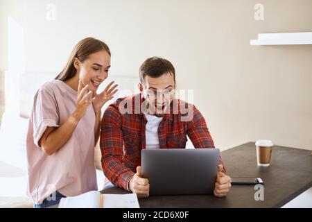 Attraktive verspielte Frau lässig gekleidet stehen im Schlafzimmer vor dem Laptop-Computer mit erhobenen Händen und geöffneten Mund, zufrieden mit ausgezeichnet Stockfoto