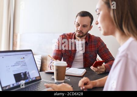 Aufmerksamer einverstimmter junger Mann, der gegenüber einer Brünette sitzt, mit besorgtem Gesicht auf einen Laptop schaut und auf Informationen wartet, die dow schreiben