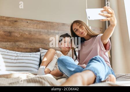 Charmante junge Mutter und kleine hübsche Mädchen verbringen Morgenzeit zusammen, Selfie, Blick direkt auf Handy-Kamera mit freudigen expressio Stockfoto