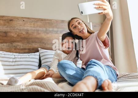 Zwei schöne Mädchen mit Spaß im Schlafzimmer, attraktive Frau macht Foto mit Handy, zeigt Zunge voller Freude, Hintergrund von kleinen Kissen, mor Stockfoto