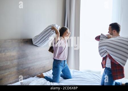 Charmante, überfröhliche Liebhaber, die Spaß zu Hause haben, im Schlafzimmer spielen, fröhlich mit Kissen kämpfen, wegschauen, breit lächeln, glücklichen Ausdruck bringen Stockfoto