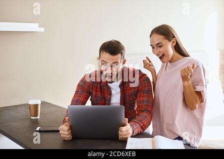 Fröhlicher Mann trägt roten Shitr und weißes T-Shirt, in runden Brillen, sitzt am Tisch, hält Computer mit beiden Händen, starrt ar Bildschirm, schreit laut, Stockfoto