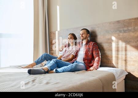 Glücklich aufgeregt Paar verbringen Freizeit zu Hause, sitzen in hell beleuchteten Schlafzimmer, Fernsehen, wegschauen, posieren auf dem Hintergrund des großen Lichts wi Stockfoto