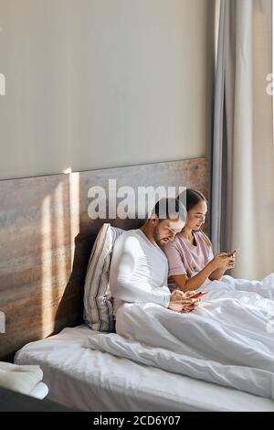 Junge, angenehme Liebhaber im Pyjama, glückliche Zeit im Hotelzimmer, Sonne scheint durch das große Fenster, SMS mit Freunden über Handys, Blick weg Stockfoto