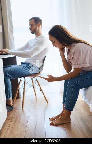 Seitenaufnahme einer müden Frau mit langen Haaren, barfuß im Schlafzimmer sitzend, Hauskleidung tragend, Handy in der Hand mit ruhigem Gesicht anblickend, wartet auf Stockfoto