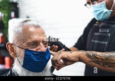 Männliche Friseur schneiden Haare zu Hipster Senior Client während des Tragens Gesicht chirurgische Maske - Junge Friseurin arbeitet in Friseursalon während corona-Virus Stockfoto