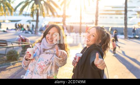 Zwei junge attraktive lächelnde Frauen machen Daumen nach oben Geste und Kaffee halten, um in ihren Händen zu gehen - weibliche Freundschaft Und Spaß haben Konzept Stockfoto