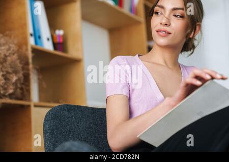 Foto von einer schönen Frau, die Buch liest, während sie darauf sitzt Sessel im hellen Büro Stockfoto