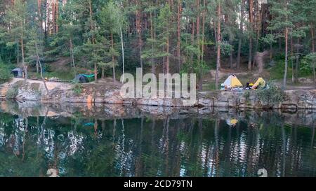 Camping am Ufer des überfluteten Granitsteinbruchs. Touristen richten Zelte für die Nacht am Ufer eines Sees in einem Kiefernwald auf Stockfoto