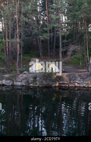 Schöne Landschaft, Camping und Zelte unter einem Kiefernwald am Rande eines überfluteten Granitbruchs, vertikale Aufnahme Stockfoto