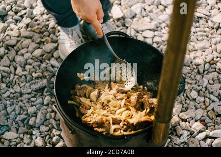 Der Koch kocht Pilaf im Kessel auf einem offenen Feuer draußen und rührt mit einer großen Pfanne. Stockfoto