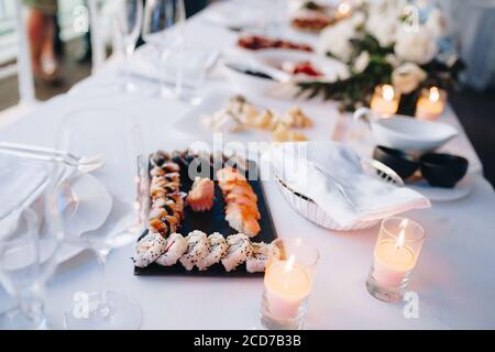 Schwarzer Teller zum Servieren von Sushi und Brötchen auf einer weißen Tischdecke auf einem Tisch mit Gläsern, Handtüchern und brennenden Kerzen. Stockfoto