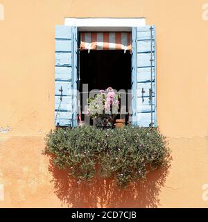Fenster mit blauen hölzernen Fensterläden und gestreiften Markise auf einer orange bemalten Wand, auf der Fensterbank, ein Topf mit Blumen, auf der Insel Burano, Venic Stockfoto