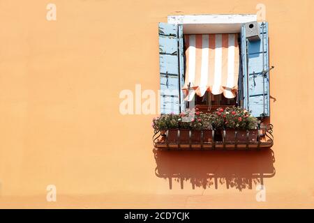 Fenster mit blauen hölzernen Fensterläden und gestreiften Markise auf einer orange bemalten Wand, auf der Fensterbank, ein Topf mit Blumen, auf der Insel Burano, Venic Stockfoto