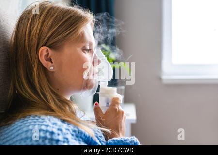 Portrait einer jungen blonden Frau, die zu Hause mit Dampfnebelvernebler inhaliert. Asthma, Grippe, Gesundheitskonzept. Stockfoto