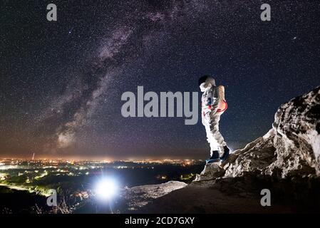 Seitenansicht des tapferen Weltraumreisenden, der auf einem felsigen Berg unter dem Nachthimmel mit Sternen steht. Mission Spezialist Astronaut im Raumanzug genießen Blick auf Nacht Stadt. Konzept der Milchstraße und Raumfahrt. Stockfoto