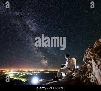 Spaceman Reisender sitzt auf felsigen Berg unter fantastischen Sternenhimmel. Raumfahrt Reisende genießen schöne Aussicht auf Milchstraße und Nacht Stadt im Tal. Konzept der Kosmonauten und Raumfahrt. Stockfoto