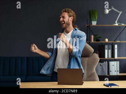 Junge gutaussehende langhaarige lockige Mann arbeitet im Büro mit Laptop und zum Ausdruck Positivität. Glücklicher Geschäftsmann bekam positive News.rewarded, neu Stockfoto