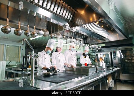 Köche in Schutzmasken und Handschuhen bereiten Essen in der Küche eines Restaurants oder Hotels zu. Stockfoto