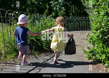 Zwei kleine Kinder halten sich die Hände und gehen einen Fußweg in Sommerkleidung und Sonnenschein Stockfoto