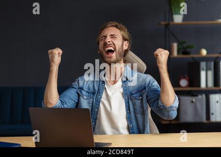 Junge gutaussehende langhaarige lockige Mann arbeitet im Büro mit Laptop und zum Ausdruck Positivität. Glücklicher Geschäftsmann bekam positive News.rewarded, neu Stockfoto