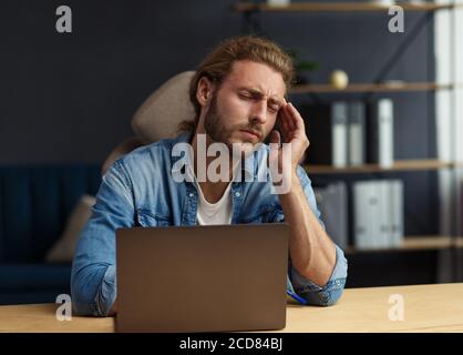 Burnout- und Überarbeitungs-Konzept. Müde Geschäftsmann Überstunden im Büro mit Laptop. Gestresster junger schöner lockiger Mann mit langen Haaren suchen Stockfoto