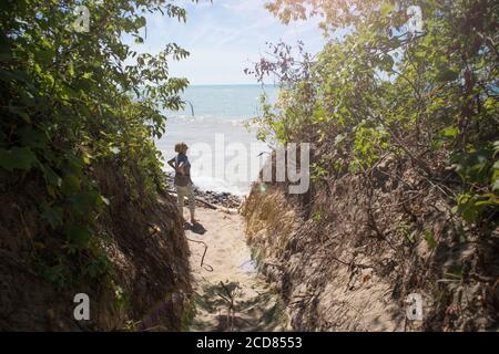 Am Ende des schmalen Durchgangs, der zum Seestrand führt, blickt eine reife Frau allein auf die Wolken. Stockfoto