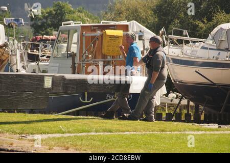 Zwei Männer, die beide helfen, das Schleusentor zu öffnen, indem sie den Balancebalken bei Schleuse fünf in Cairnbaan am Crinan Canal, Schottland mit Booten hinter sich schieben Stockfoto