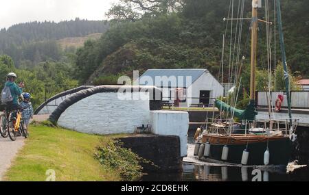 Eine Mutter und ein kleiner Sohn beobachten eine Holzyacht, die durch die geöffnete Bellanoch Bridge auf dem Crinan Canal, Argyll, Schottland, fährt Stockfoto