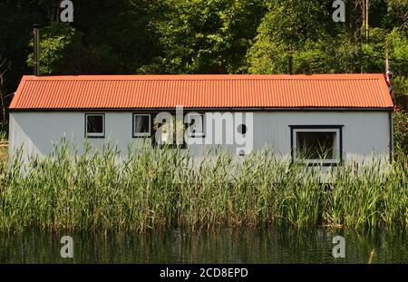 Ein Bungalow mit einem roten Metalldach am Ufer des Crinan Canal, Argyll, Schottland in der Nähe der Bellanoch Road Swing Bridge mit Schilf und Reflexionen Stockfoto