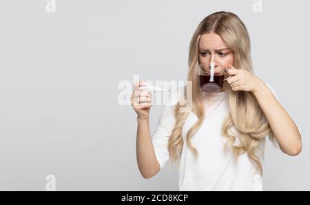 Frau in T-Shirt hält Tasse heißen Tee, erwischt kalt und Blick auf Thermometer, isoliert auf grauem Hintergrund. Grippesaison. Stockfoto